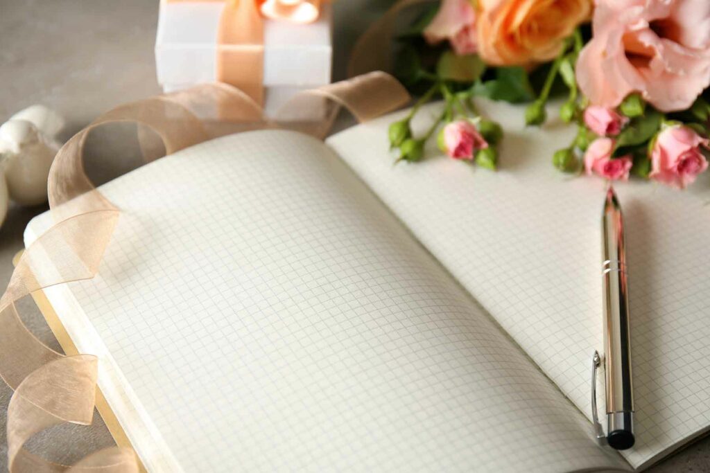 A blank notebook for writing wedding vows surrounded by a ring box, florals and a silver pen