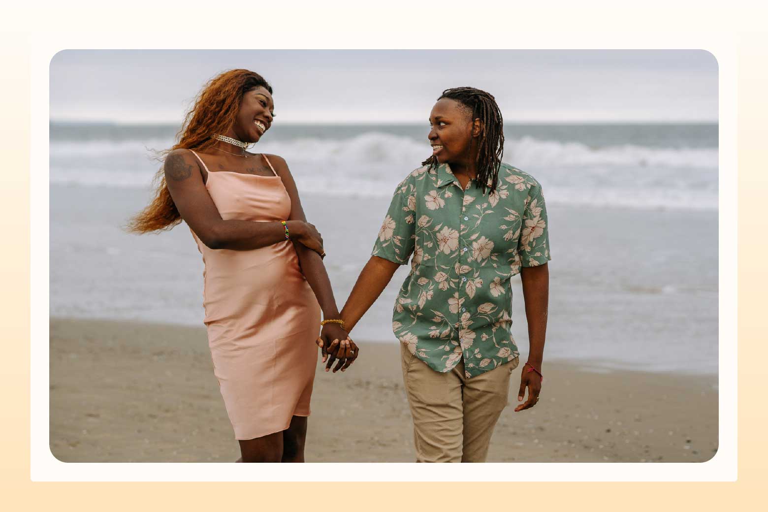 Lesbian couple walking on beach while holding hands and smiling at each other