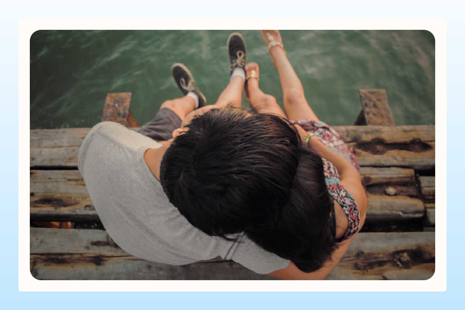 Overhead shot of a couple sitting side by side on a pier, his head leaning onto hers, with their legs dangling over the water