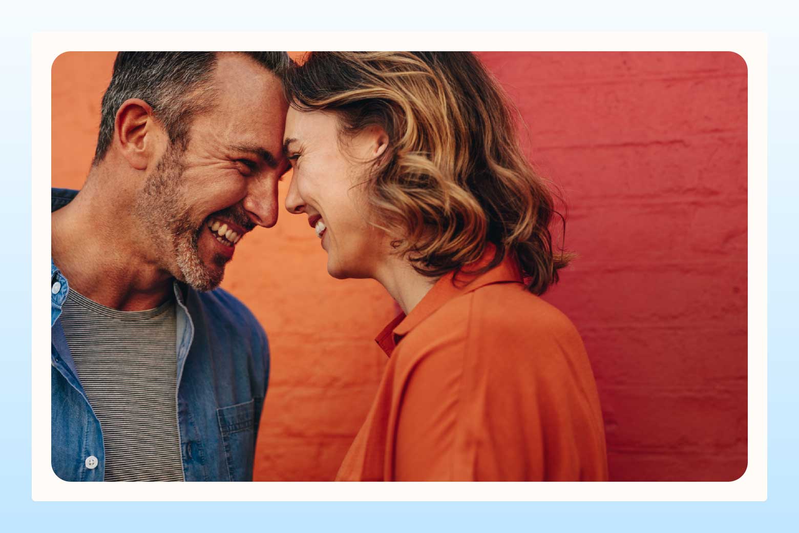 Smiling man and woman lean their foreheads against one another, standing in front of a bright orange and red wall