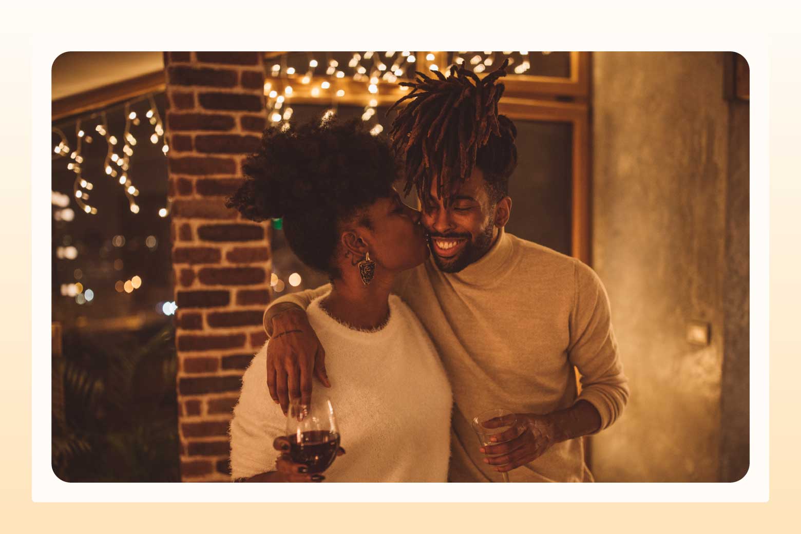 Woman kisses man whose arm is around her shoulders, with twinkling white lights and a brick column behind them