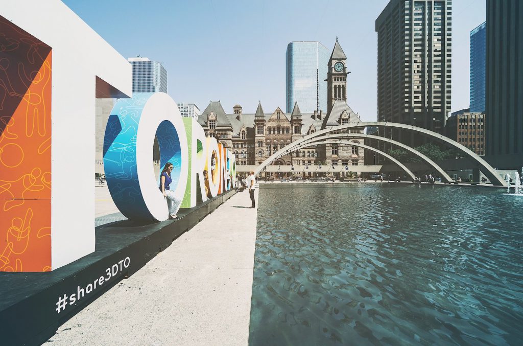 view of nathan phillips square in toronto featuring water, arches, a large toronto sign, and buildings in the background