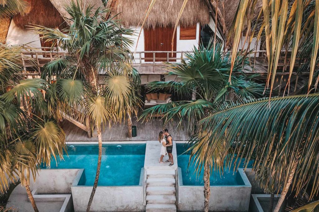 A couple embracing on their honeymoon on a bridge above a private pool, surrounded by palm trees and villas in the background