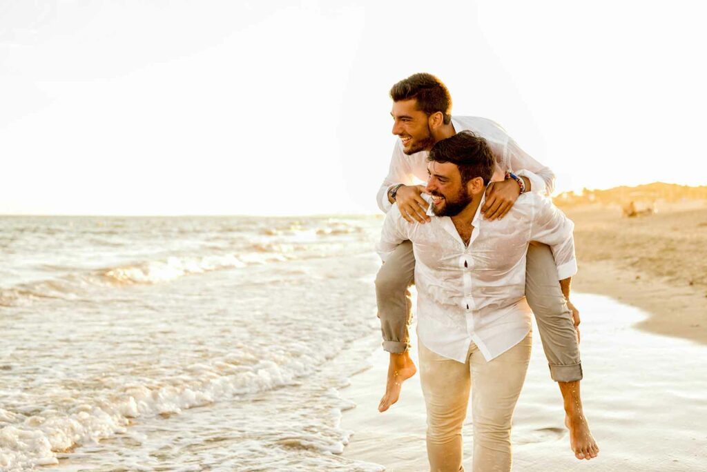 A man carrying his partner on his back during their beach honeymoon walking along the shore