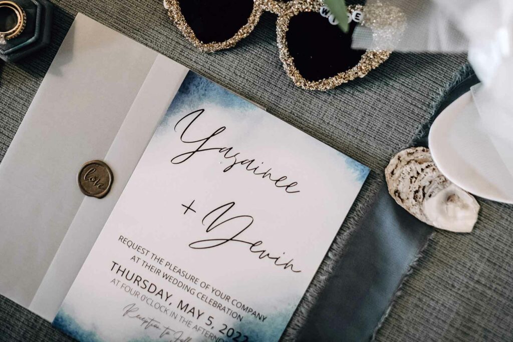 A wedding invitation with watercolor edges surrounded by sunglasses, wedding rings, and a seashell