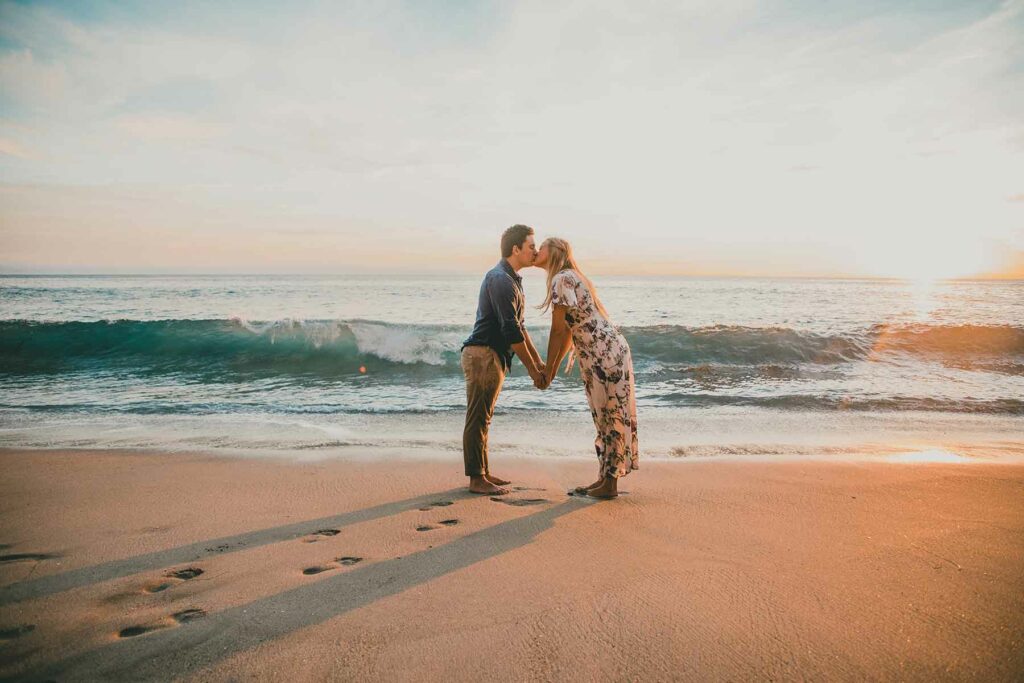 What to Wear in Engagement Photos: Couple kissing on a beach holding hands with a man wearing a button down shirt and dress pants and a woman wearing a floral dress
