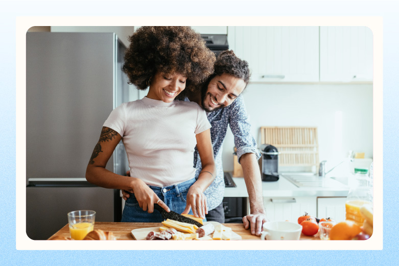 Couple standing in kitchen, man with arms around woman as she slices meats and cheese