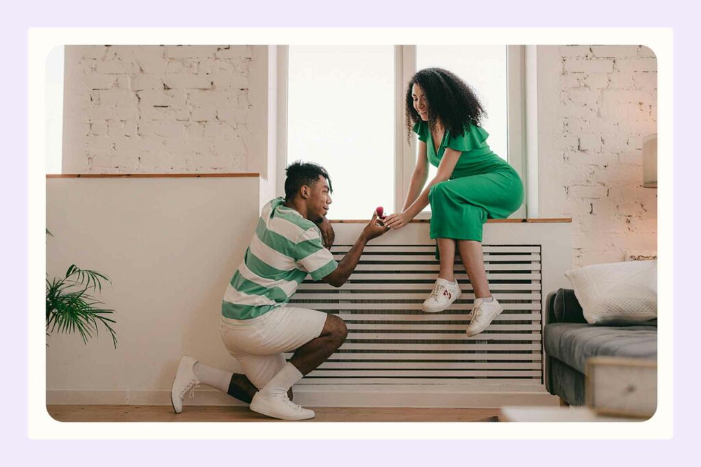 A kneeling man holding an open ring box in a bedroom while a woman perches on a windowsill