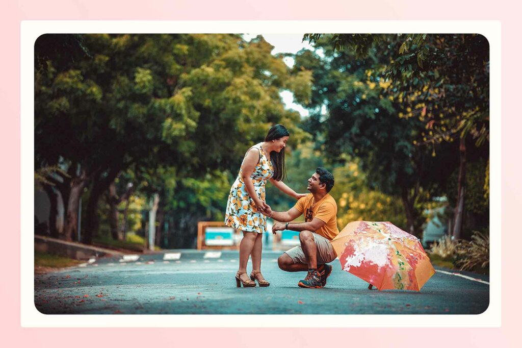 A man kneeling on a small town street lined with trees, about to propose to a woman holding his hand