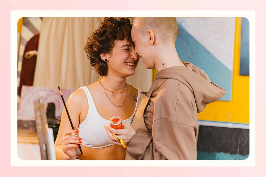 A couple smiling and embracing with their eyes clothes. One is holding a paintbrush and the other is holding a paintbrush and paint bottle