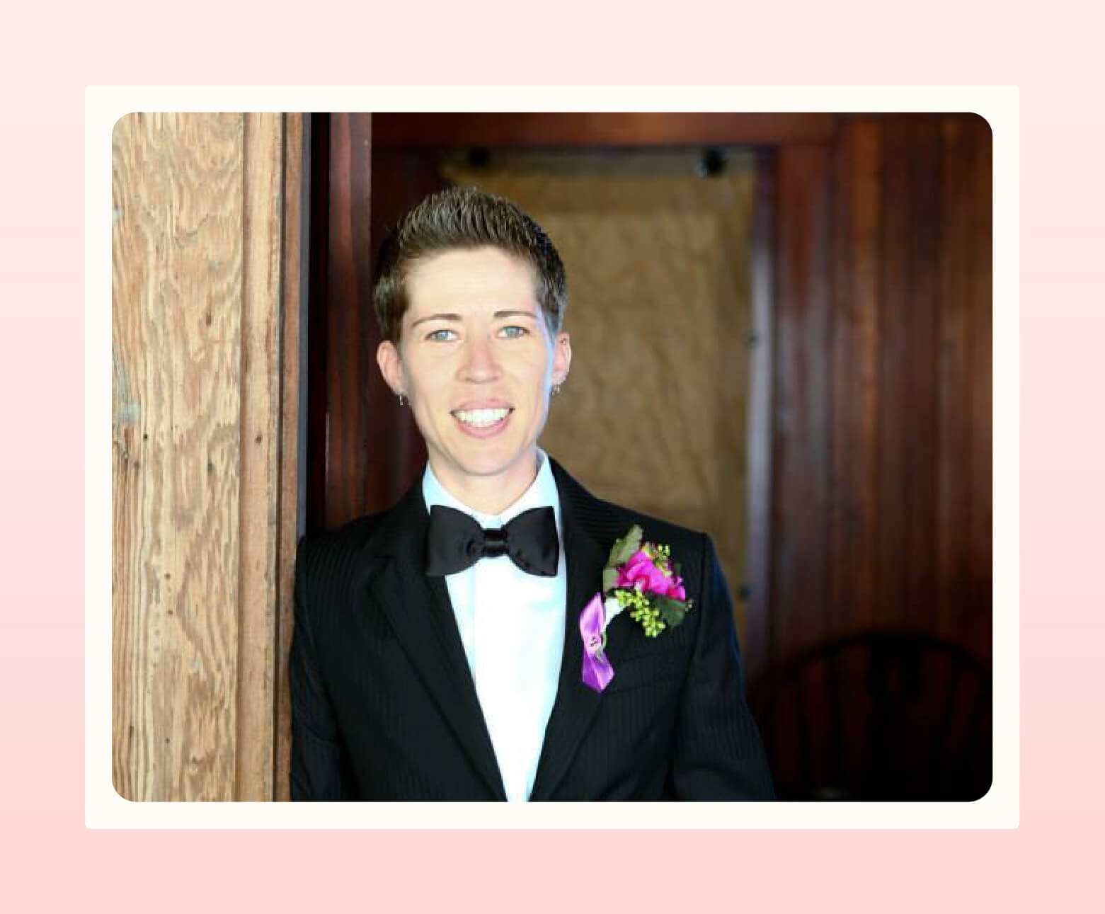 Woman leaning against doorway wearing a black suit with black bow tie and purple and green boutonniere