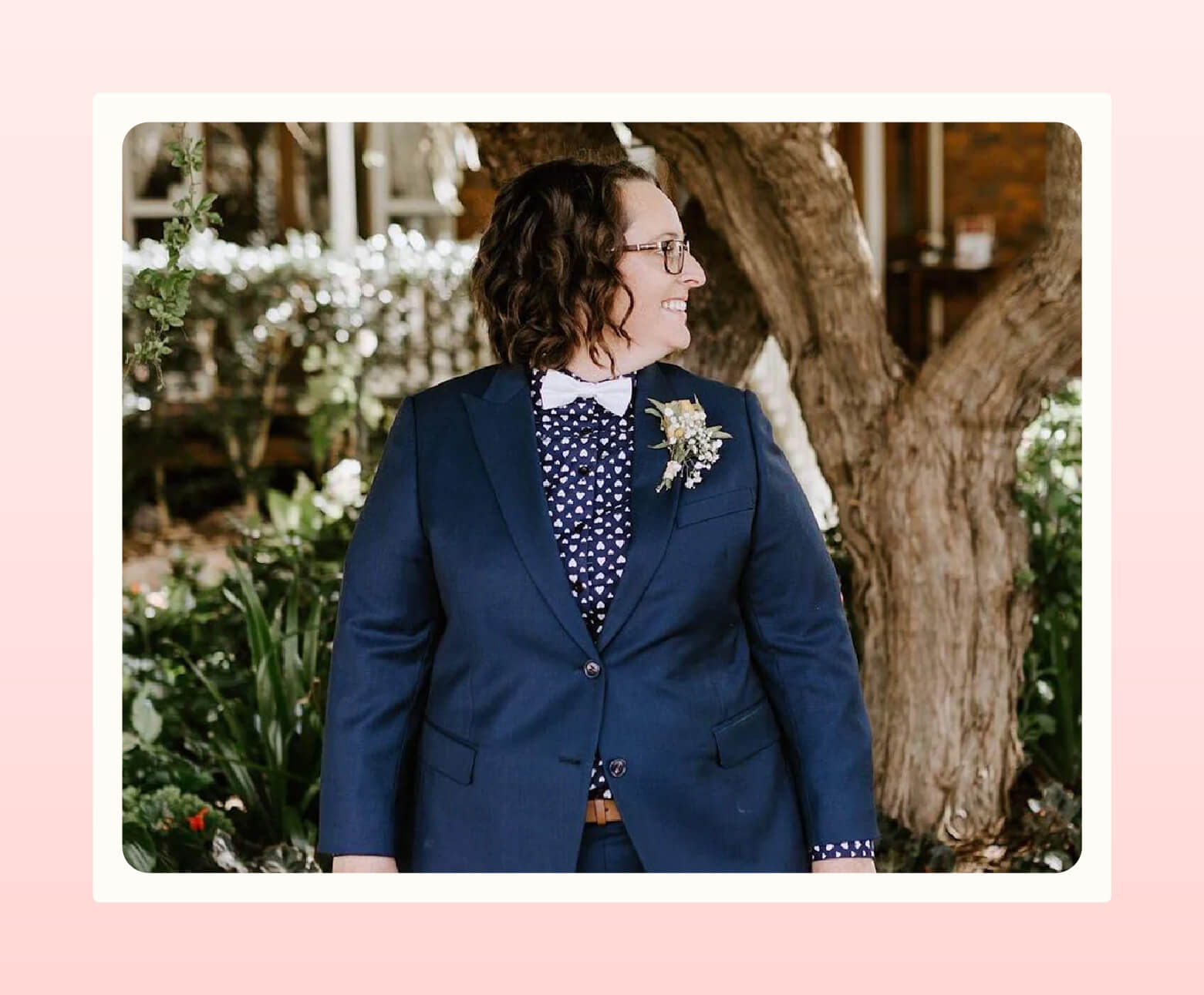 Person in navy blue suit from Shane Ave. with a white bow tie and a boutonniere looks to their left with a smile on their face